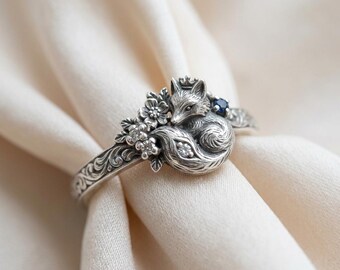 May include: A silver ring featuring a sculpted fox wearing a small crown, accented with floral details, clear stones, and a single dark blue stone. The band is decorated with intricate scrollwork engravings along the sides of the metal surface.