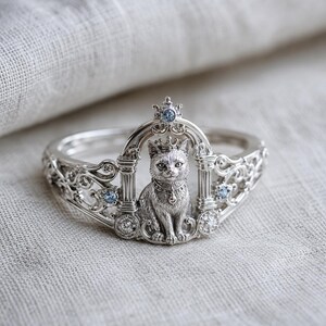 May include: A silver ring featuring a detailed cat design. The ring has a cat figure wearing a crown, set within an ornate archway. The ring is embellished with small blue gemstones and intricate filigree details.