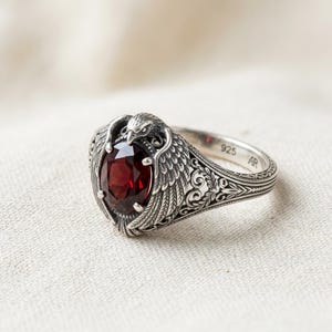 Könnte beinhalten: Ein silberner Ring mit einem ovalen, tiefroten Edelstein. Er zeigt ein Adlerdesign mit detaillierten Flügeln und Kopf sowie aufwendige Filigranmuster auf dem Band. Der Ring ist mit "925 AR" gestempelt.
