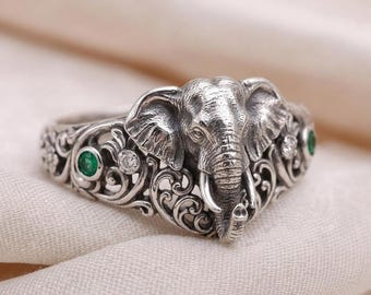 May include: A silver ring featuring a detailed elephant head centerpiece with prominent tusks. The band is decorated with intricate scrollwork, two small round green gemstones, and two clear round stones set on either side of the elephant.
