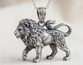 May include: A silver lion pendant with a detailed mane and tail, adorned with red and blue gemstones. The pendant is suspended from a delicate silver chain, showcasing intricate scrollwork and a regal design.