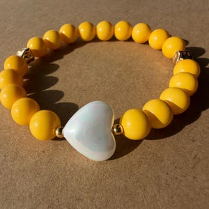 May include: A yellow beaded bracelet with a pearlescent white heart-shaped charm. The bracelet features round yellow beads and gold-colored accents. The heart charm is centered on the bracelet.
