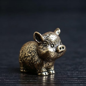 Könnte beinhalten: Kleine, detaillierte Bronzefigur eines Schweins. Das Schwein ist in sitzender Position dargestellt, mit aufgestellten Ohren und sichtbarer Schnauze. Die Figur hat eine strukturierte Oberfläche und steht vor einem dunklen Hintergrund.