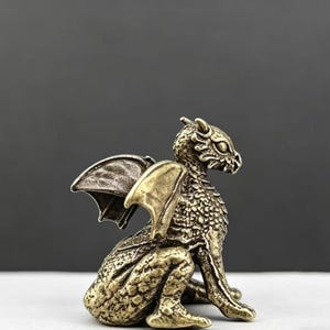 May include: A detailed bronze-colored dragon figurine with textured scales, wings, and a fierce expression. The dragon is in a seated position, showcasing its intricate design and craftsmanship. The wings are spread, and the overall aesthetic is fantasy-themed.
