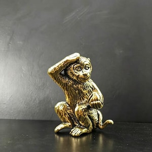 Puede incluir: Pequeña figura de mono dorada, sentado, con una mano en la cabeza. El mono tiene detalles finos y una superficie texturizada, sugiriendo bronce o latón. La figura está sobre una superficie oscura.