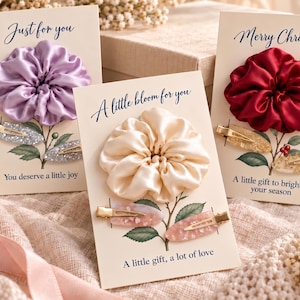 May include: Three hair accessory sets on display cards. Each set includes a satin scrunchie and two hair clips. The cards feature floral illustrations and text: Just for you, A little bloom for you, and Merry Christmas, with additional phrases.