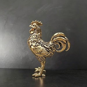 May include: A detailed, golden-colored rooster figurine. The sculpture features intricate feather detailing, a prominent comb, and a curved tail. The rooster stands on two legs, with its head held high. The background is a dark gray.