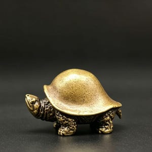 Puede incluir: Pequeña figura de tortuga de latón detallada. La tortuga tiene un caparazón y un cuerpo texturizados, con patas visibles y una cara detallada. La figura es de color oro antiguo cálido.