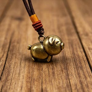 May include: A bronze-colored pig pendant on a brown and orange cord necklace. The pig charm has detailed features, including a snout, ears, and a curled tail. The necklace is displayed on a wooden surface.