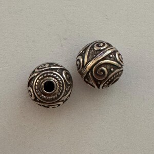 May include: Two spherical metal beads featuring a dark oxidized finish and a raised, polished silver-toned swirl pattern. Each bead has a central hole for threading, with intricate textured detailing surrounding the opening and across the surface.