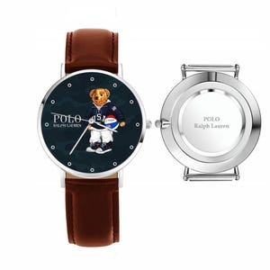 May include: A wristwatch with a brown leather band and a silver-colored case. The watch face features a navy blue background with a bear wearing a jacket and holding a ball. The watch face also displays the text "POLO Ralph Lauren".