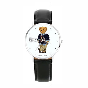 Puede incluir: Un reloj de pulsera con una correa de cuero negro y una esfera blanca. La esfera del reloj presenta un oso de dibujos animados con un suéter azul marino y pantalones color caqui. La esfera del reloj también tiene el texto "POLO RALPH LAUREN" y "SWISS MADE".