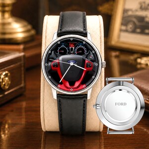 May include: A silver wristwatch with a black leather band. The watch face features a steering wheel design with red accents and the Ford logo. A separate watch back with the word "FORD" is also visible.