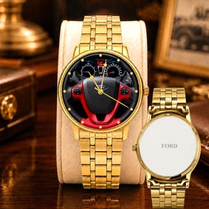 May include: Gold-toned wristwatch with a black and red steering wheel design on the face. The watch has a matching gold band and a white backing with the word "FORD". The watch is displayed on a beige stand.