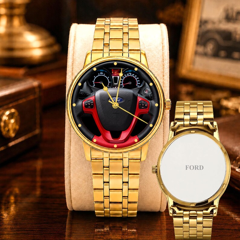 May include: Gold-toned wristwatch with a black and red steering wheel design on the face. The watch has a matching gold band and a white backing with the word "FORD". The watch is displayed on a beige stand.