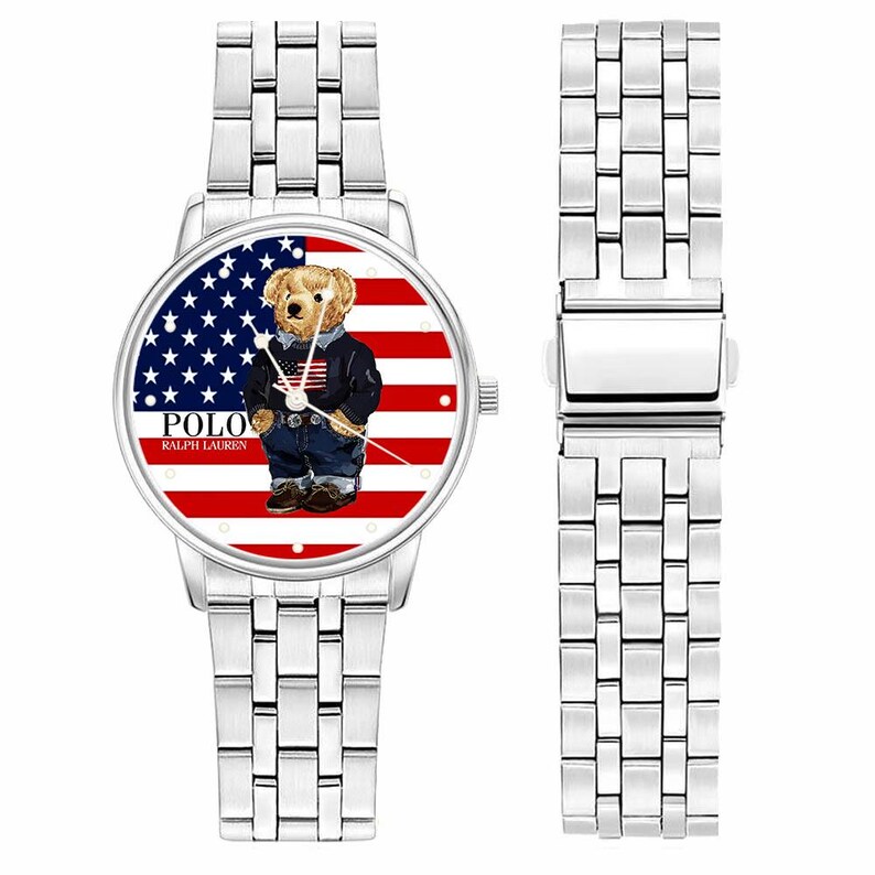 May include: A silver wristwatch with a round face featuring a bear wearing a jacket and jeans against a backdrop of the American flag. The watch face displays the text "POLO RALPH LAUREN". The watch has a silver band.