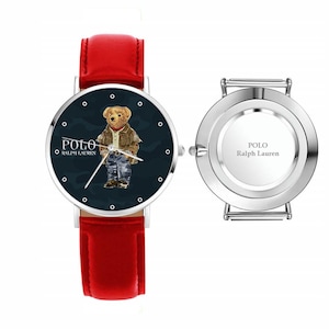Op de afbeelding: Een horloge met rode leren band en een marineblauwe wijzerplaat met een beer die een jas en jeans draagt. Op de wijzerplaat staan de woorden "POLO" en "RALPH LAUREN". Het horloge wordt getoond naast de achterkant van het horloge.