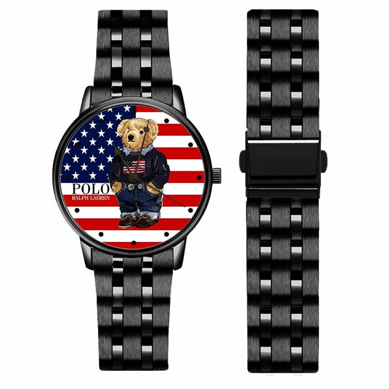 May include: Black wristwatch with a round face featuring a bear wearing a jacket and jeans in front of an American flag design. The watch has a black metal band and the text "POLO RALPH LAUREN".