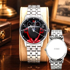 May include: Silver wristwatch with a black and red steering wheel design on the watch face. The watch has a silver band and is displayed on a tan watch stand. A second watch with the word "FORD" is also visible.