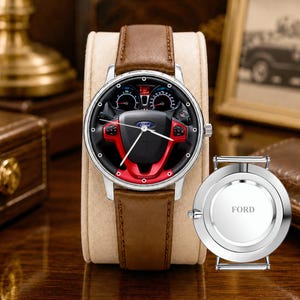 May include: A silver wristwatch with a brown leather band. The watch face features a black and red steering wheel design with the Ford logo. The watch is displayed on a beige stand, with a silver back cover that says "FORD".