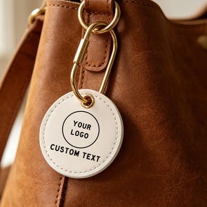 May include: A round white leather tag with black text reading YOUR LOGO and CUSTOM TEXT, attached by a gold-toned metal carabiner to a brown leather handbag. The tag features visible stitching along the circular edge and a gold-toned eyelet.