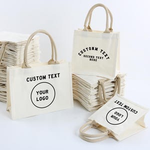 May include: A collection of off-white canvas tote bags with beige handles. Some bags are stacked, while others stand upright. The bags feature black printed text that reads CUSTOM TEXT, YOUR LOGO, and CUSTOMM TEXT SECOND TEXT HERE.