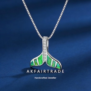 Puede incluir: Un collar plateado con un colgante en forma de cola de ballena. El colgante es verde y blanco con un diseño texturizado y está adornado con pequeñas gemas transparentes. El collar tiene el texto "AKFAIRTRADE" y "Handcrafted Jeweller".