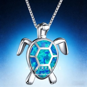 Op de afbeelding: Een zilverkleurige schildpadhangerketting met een blauw-groene opaalachtige schaal. De hanger is bevestigd aan een fijne ketting. De tekst "AKFAIRTRADE Handcrafted Jewel" is zichtbaar op de hanger.