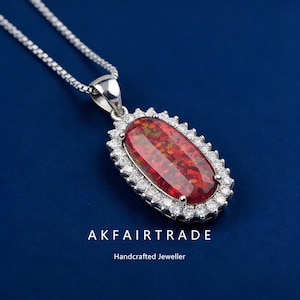 Peut inclure: Un collier en argent avec un pendentif orné d'une grande pierre précieuse rouge ovale entourée de petites pierres claires. Le pendentif est suspendu à une fine chaîne en argent. Le texte "AKFAIRTRADE Handcrafted Jeweller" est en bas.