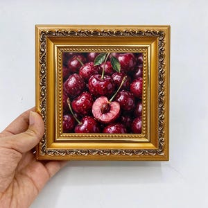 May include: A square painting of dark red cherries with water droplets, featuring one halved cherry showing the pit and a green leaf, encased in an ornate, metallic gold-coloured frame with intricate embossed detailing along the edges.