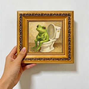 Puede incluir: Una pintura al óleo enmarcada que muestra una rana verde con los brazos cruzados sentada en un inodoro blanco. La obra está dentro de un marco dorado ornamentado con intrincadas tallas decorativas en los bordes. El texto AKFAIRTRADE es visible.