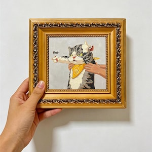May include: A square painting in an ornate gold frame featuring a gray and white tabby cat wearing a yellow bandana. The cat is depicted with a grumpy expression, pointing one paw to the side and raising the other, with the text "Bui~" visible.
