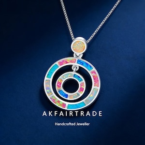 May include: A silver necklace with a pendant featuring concentric circles inlaid with colorful opal stones. The pendant hangs from a delicate chain. The text "AKFAIRTRADE Handcrafted Jeweller" is at the bottom.