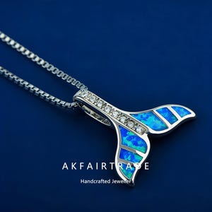 May include: A silver-toned pendant necklace featuring a whale tail design. The tail is inlaid with blue and green opal-like material and accented with clear gemstones. The necklace has a delicate chain and is set against a deep blue background. The text "AKFAIRTRADE Handcrafted Jeweller" is at the bottom.