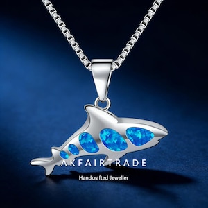May include: A silver pendant necklace with an orca whale design. The whale has blue opal-like inlays and hangs from a fine chain. The text "AKFAIRTRADE Handcrafted Jeweller" is visible. The background is a deep blue.