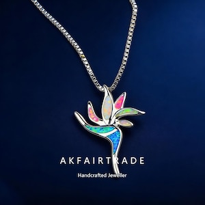 Könnte beinhalten: Eine silberfarbene Halskette mit einem Anhänger in Form einer Paradiesvogelblume. Der Anhänger hat ein Mosaik aus schillernden Farben, darunter Rosa, Blau, Grün und Weiß. Der Text "AKFAIRTRADE Handcrafted Jeweller" ist zu sehen.
