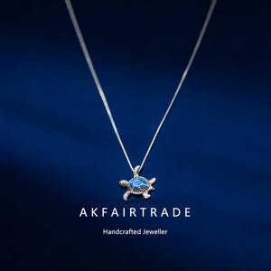 Op de afbeelding: Een zilveren ketting met een schildpadhanger. De hanger heeft een blauwe emaille schild, zilveren details en kleine heldere stenen. De ketting hangt tegen een donkerblauwe achtergrond. De tekst "AKFAIRTRADE" en "Handcrafted Jeweller" staan eronder.