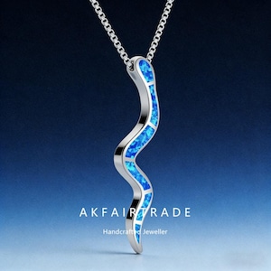 May include: A silver necklace featuring a flowing, serpentine pendant. The pendant is set with vibrant blue, rectangular inlays. The necklace has a fine chain. The text "AKFAIRTRADE Handcrafted Jeweller" is displayed at the bottom.
