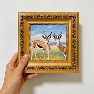 May include: An oil painting of two springbok antelopes standing in a grassy field with mountains in the background, encased in a square, ornate, metallic gold frame with a decorative embossed border and a smooth inner trim.