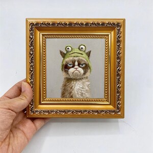 May include: A square oil painting in an ornate gold frame featuring a portrait of a grumpy-looking cat wearing a green frog-shaped hat with large eyes. The cat has brown and white fur and a stern facial expression. The text AKFAIRTRADE is visible.