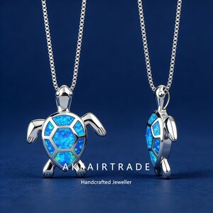 Könnte beinhalten: Eine silberfarbene Schildkröten-Anhänger-Halskette mit blauem Opal-Inlay. Der Anhänger hängt an einer silbernen Kette. Der Text "AK AIRTRADE" und "Handcrafted Jeweller" ist ebenfalls zu sehen. Der Hintergrund ist einfarbig blau.