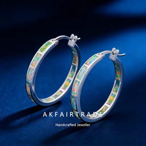 Può includere: Un paio di orecchini a cerchio in argento con intarsi di opale colorati. Gli orecchini hanno un design rettangolare con una varietà di colori, tra cui verde, giallo e arancione. Il testo "AKFAIRTRADE" e "Handcrafted Jeweller" sono in basso.