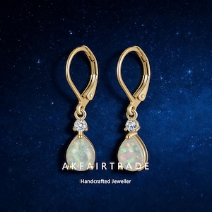 May include: A pair of gold-colored leverback earrings with teardrop-shaped opal pendants. Each earring features a small, round, clear gemstone above the opal. The text "AKFAIRTRADE Handcrafted Jeweller" is visible at the bottom.