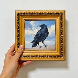 Puede incluir: Una pintura al óleo cuadrada de un cuervo negro sobre un cielo azul con nubes blancas, encerrada en un marco dorado metálico ornamentado con detalles en relieve intrincados a lo largo de los bordes. La obra presenta pinceladas visibles.