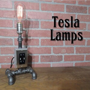 May include: A silver table lamp with a black electrical outlet on the side. The lamp is made of industrial pipe and has a vintage Edison bulb. The lamp is sitting on a wooden surface in front of a brick wall. The text "Tesla Lamps" is visible on the brick wall.