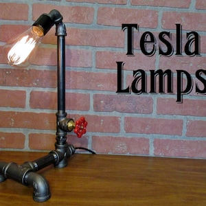 May include: A black pipe lamp with a red valve and a vintage light bulb. The lamp is sitting on a wooden table in front of a brick wall. The text "Tesla Lamps" is visible on the wall.