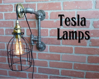 May include: A black metal cage light fixture with a gold-colored socket and a clear light bulb hangs from a black cord and pulley system attached to a gray metal pipe. The pipe is mounted to a brick wall. The text "Tesla Lamps" is written in black letters on the brick wall.