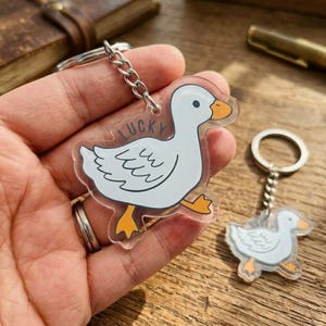 May include: Clear acrylic keychain featuring a white cartoon goose with orange feet and beak, and the word "LUCKY". The keychain is attached to a silver chain and keyring. A second, smaller goose keychain is also visible.