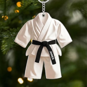 May include: A white karate uniform ornament with a black belt. The belt has the name "JAMES" printed on it. The ornament is hanging from a silver chain. The background is blurred with green and yellow lights.