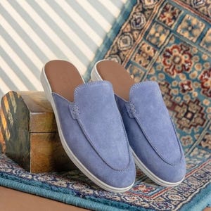Könnte beinhalten: Ein Paar Schlupfschuhe aus periwinkle-blauem Wildleder mit einer abgesteppten Mokassin-Naht an der Vorderseite, einem glatten braunen Innenfutter und einer schlichten weißen Gummisohle. Die Schuhe haben ein flaches Design mit runder Kappe.